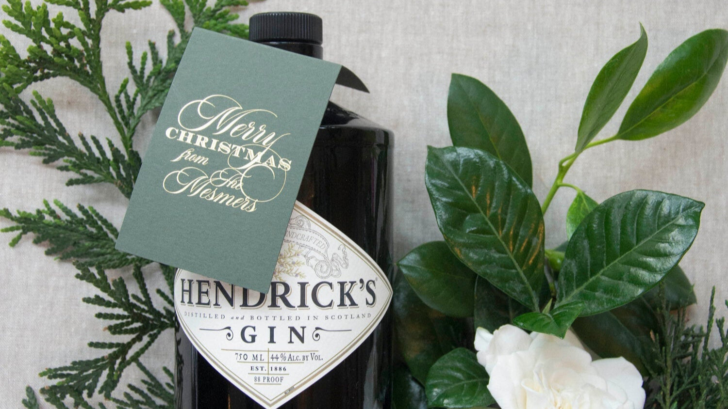 Hendrick's Gin bottle with a Christmas tag surrounded by greenery and a white flower on a textured surface.